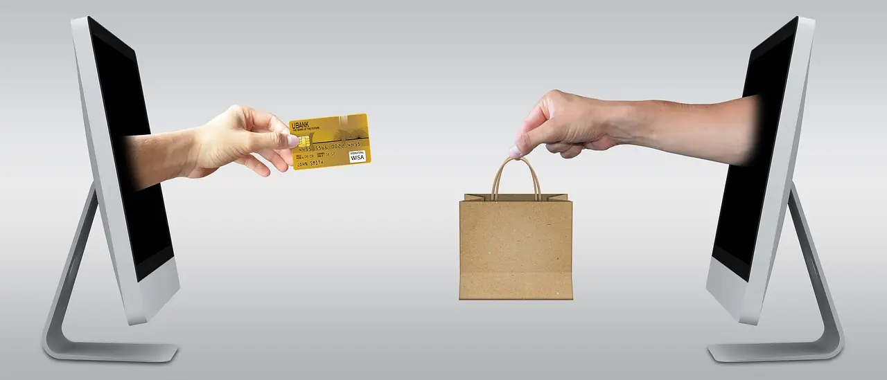 Hand with a credit card reaching toward a shopping bag between two screens, representing online shopping